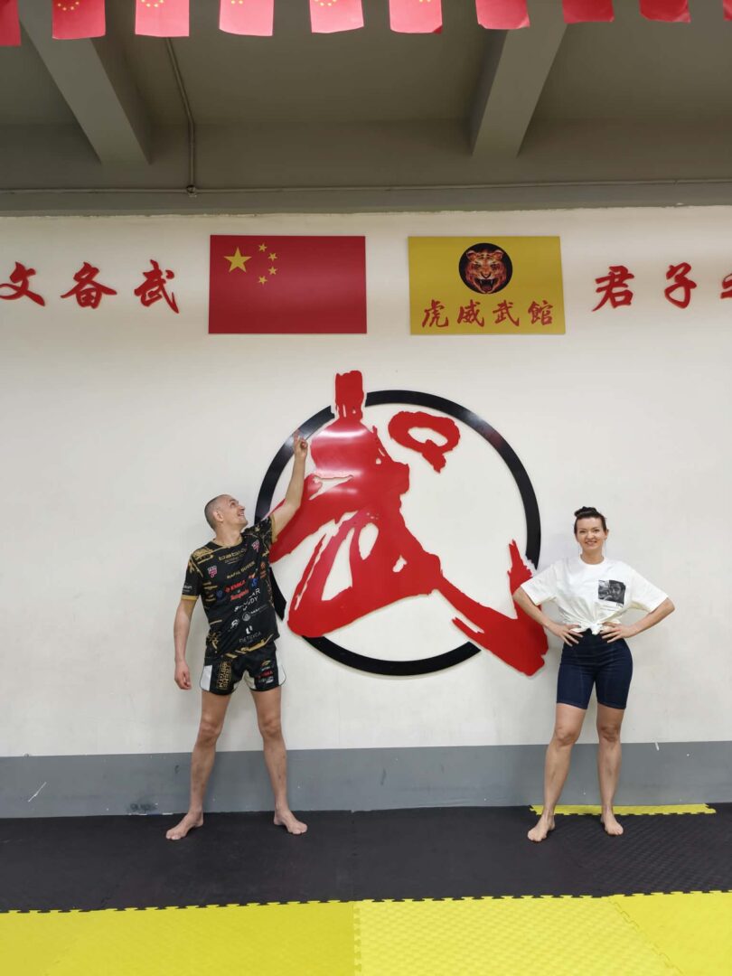 Two people in a martial arts gym pose in front of a large red and black circular emblem on the wall, with Chinese flags and banners above.