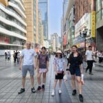 Four people stand together on a wide city street with modern buildings and shops behind them, posing for a photo.