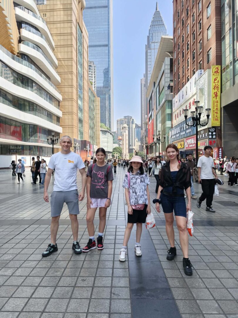Four people stand together on a wide city street with modern buildings and shops behind them, posing for a photo.
