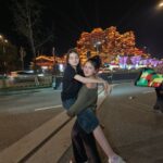Two people on a city street at night; one person lifts the other in a hug, with a brightly lit, ornate building in the background.