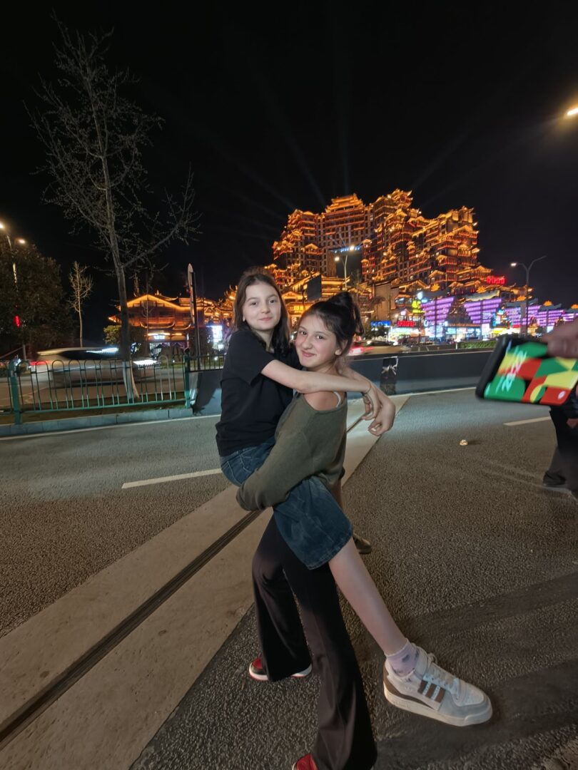Two people on a city street at night; one person lifts the other in a hug, with a brightly lit, ornate building in the background.