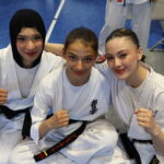 Three smiling female taekwondo athletes in white gis sit close together with their fists raised for the camera, on a blue mat.