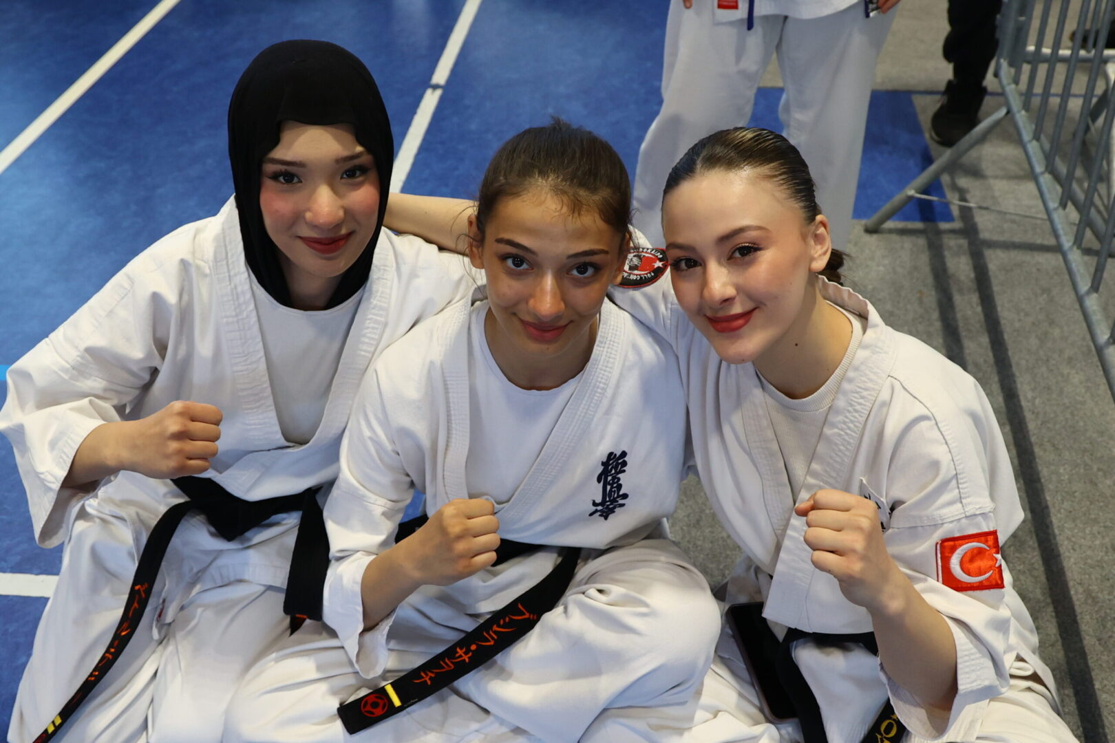 Three smiling female taekwondo athletes in white gis sit close together with their fists raised for the camera, on a blue mat.