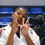 Young girl in a white karate gi makes a double peace sign and puckers her lips at the camera in a gym with blue seats in the background.