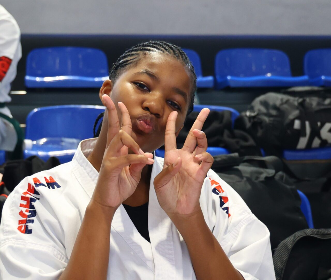Young girl in a white karate gi makes a double peace sign and puckers her lips at the camera in a gym with blue seats in the background.