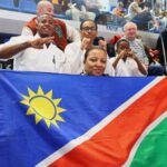 Group of karatekas posing with a Namibian flag in a sports arena, signaling victory with fists and peace signs.