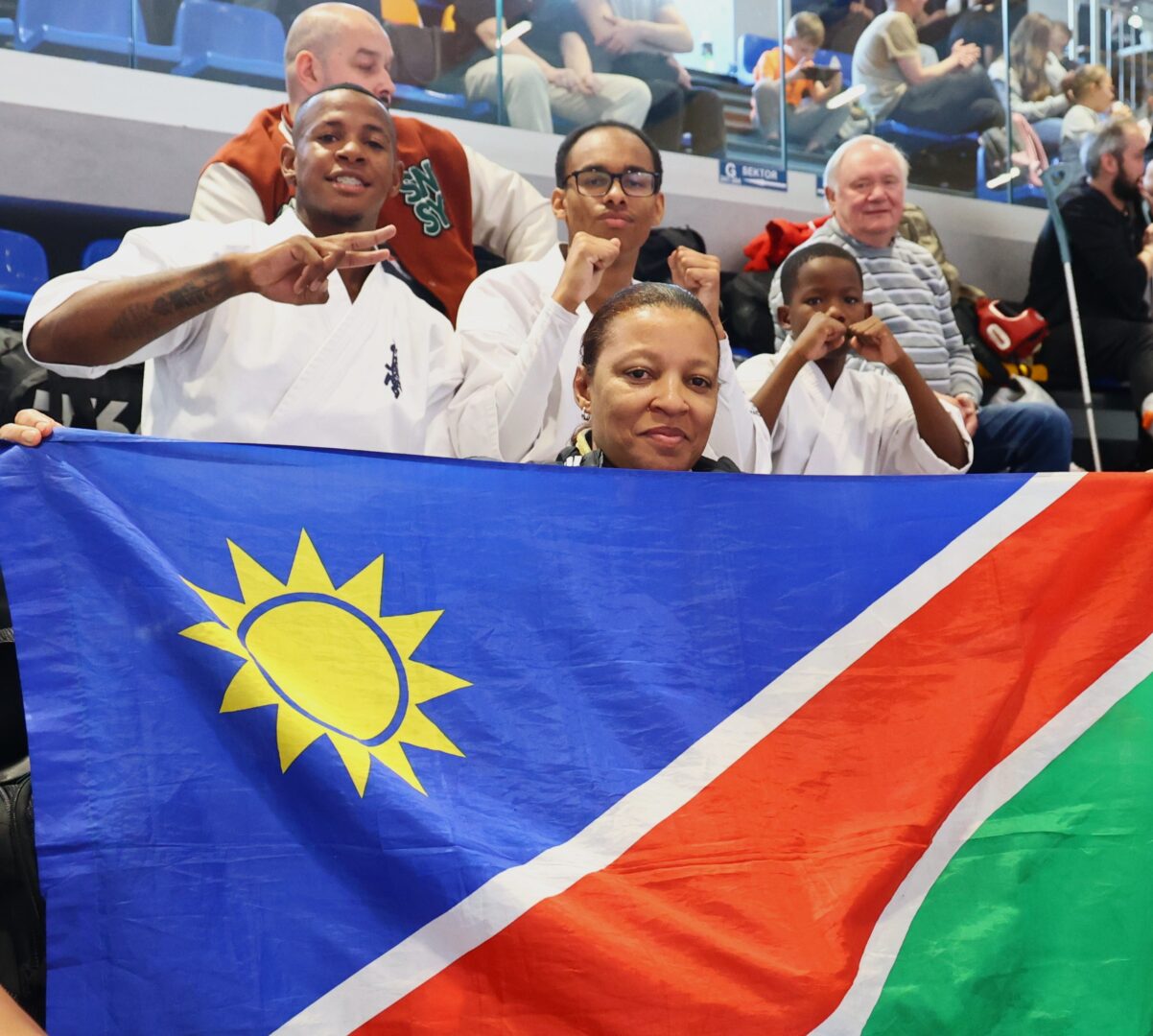 Group of karatekas posing with a Namibian flag in a sports arena, signaling victory with fists and peace signs.