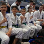 Group of young karate students in white gis with colored belts sitting in blue stadium seats, some making fists toward the camera.