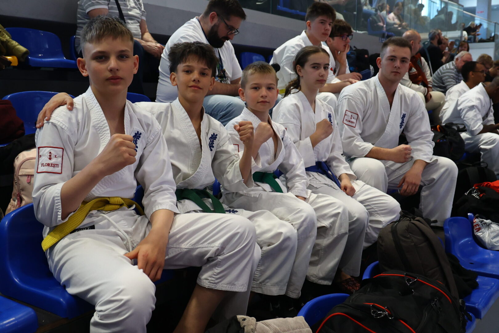 Group of young karate students in white gis with colored belts sitting in blue stadium seats, some making fists toward the camera.