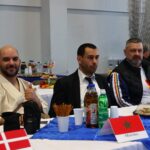 Three men sit at a long table with snacks and drinks during a formal event; Danish and Moroccan flags visible on the table.