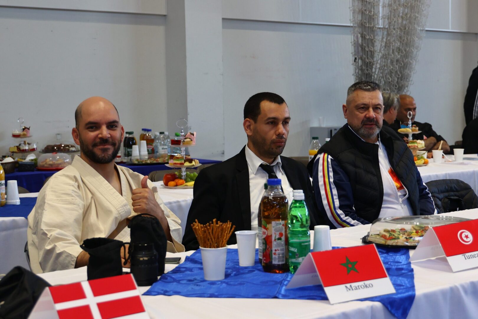Three men sit at a long table with snacks and drinks during a formal event; Danish and Moroccan flags visible on the table.
