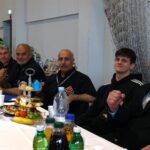 Group of men seated at a long table with snacks and drinks in a hall; some wear black uniforms, one in a suit, posing for the camera as plates of cake and fruit are visible nearby.