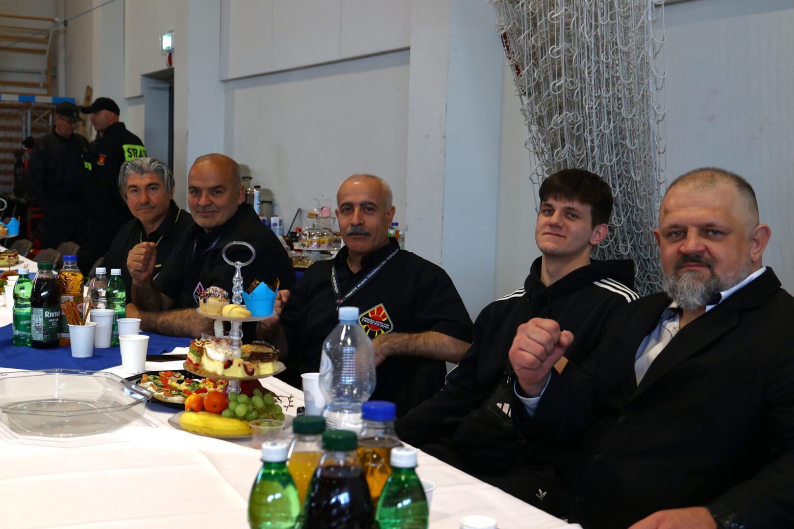 Group of men seated at a long table with snacks and drinks in a hall; some wear black uniforms, one in a suit, posing for the camera as plates of cake and fruit are visible nearby.
