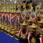 Row of gold trophies with red-and-white ribbons lined up on a blue table, ready for an awards event.