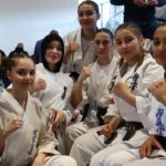 Group of young women in karate gi uniforms posing with fists raised indoors.