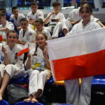 Young karate competitors in white uniforms sit on blue seats, waving small Polish flags and one holds a large Polish flag.