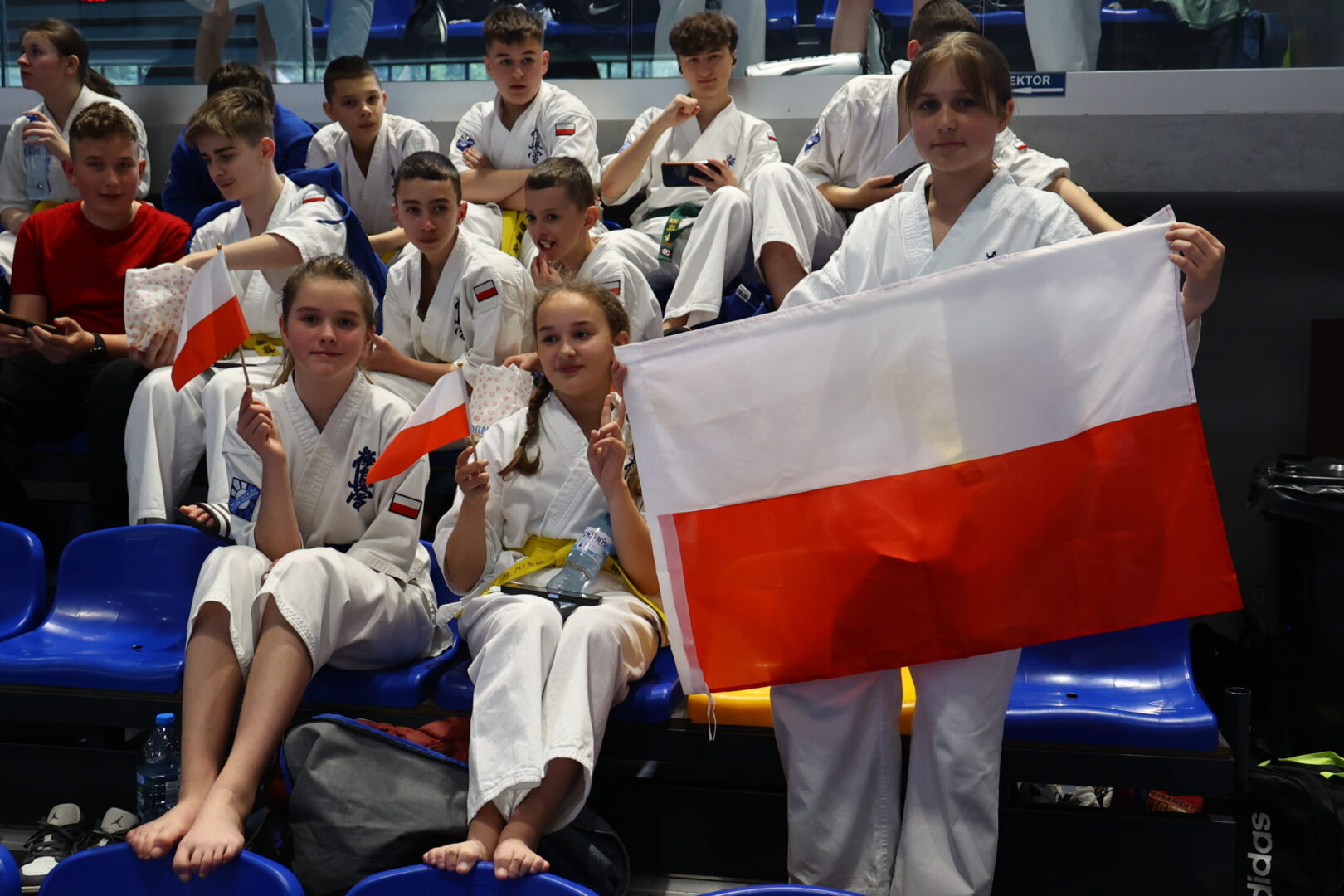 Young karate competitors in white uniforms sit on blue seats, waving small Polish flags and one holds a large Polish flag.