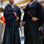 Two martial artists in navy keikogi and hakama stand in a gym, each holding a wooden bokken ready for practice.