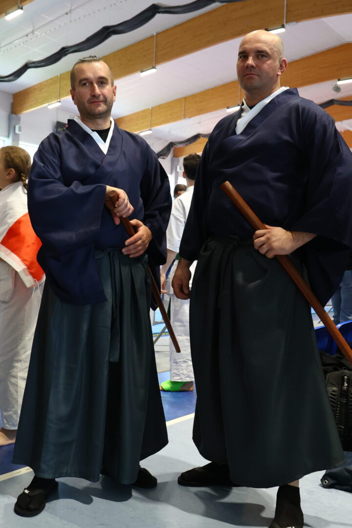 Two martial artists in navy keikogi and hakama stand in a gym, each holding a wooden bokken ready for practice.
