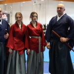 Four martial artists pose in a gym on blue mats: two men in dark blue uniforms with wooden weapons and two young women in red tops with gray skirts, crowd in the background.