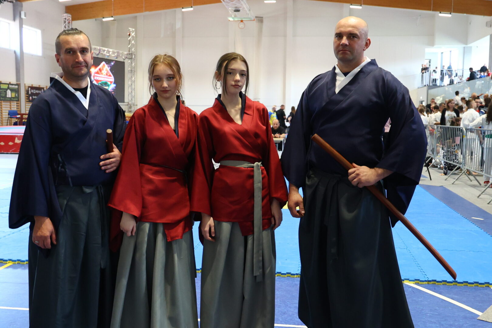 Four martial artists pose in a gym on blue mats: two men in dark blue uniforms with wooden weapons and two young women in red tops with gray skirts, crowd in the background.