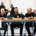 Three men in black shirts sit at a table labeled Mat D during a martial arts competition, with competition activity and people in the background.