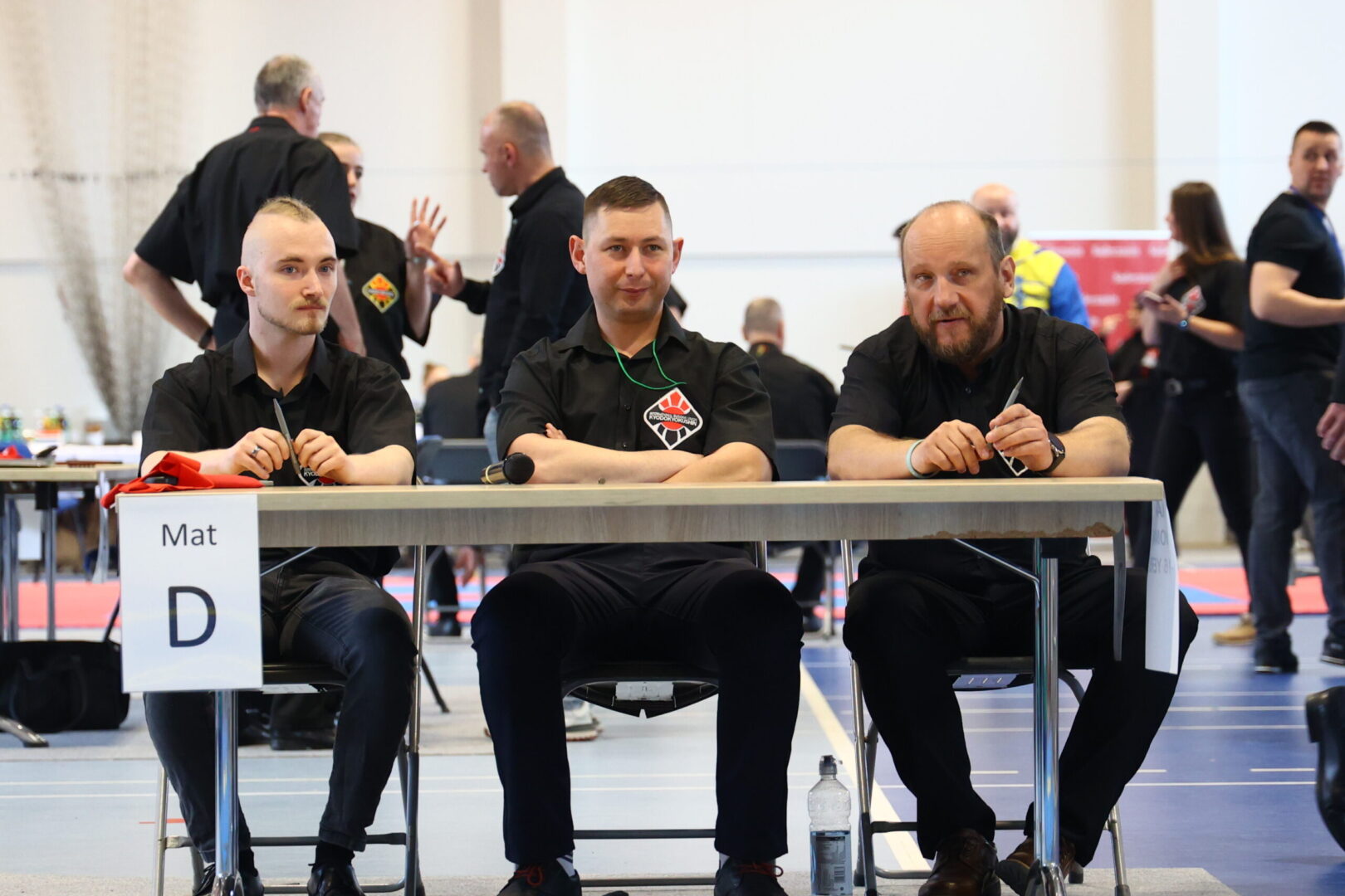 Three men in black shirts sit at a table labeled Mat D during a martial arts competition, with competition activity and people in the background.