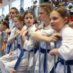 Group of young girls in white karate gi with blue belts leaning on a barrier at a martial arts event, awaiting their turn.