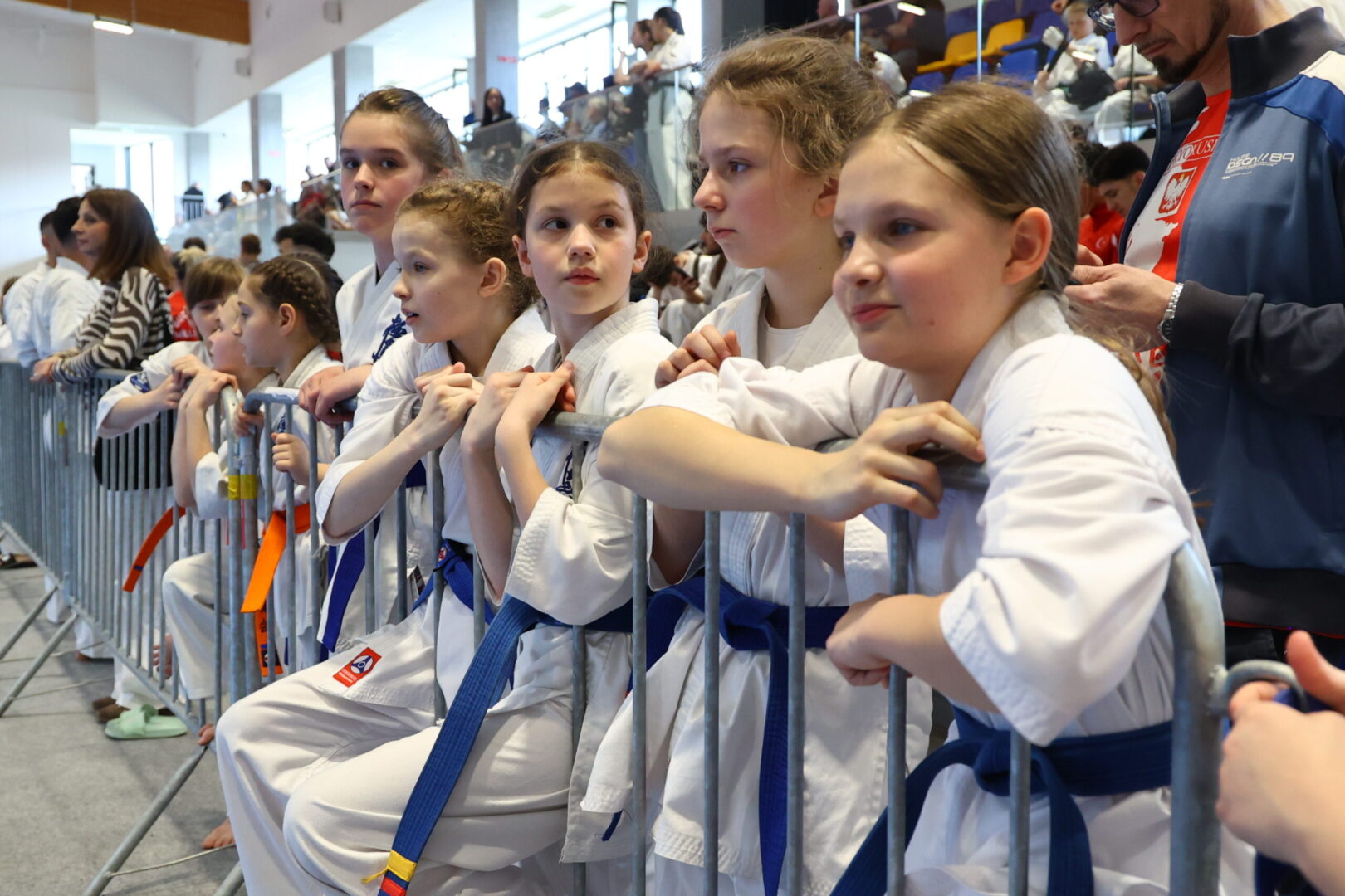 Group of young girls in white karate gi with blue belts leaning on a barrier at a martial arts event, awaiting their turn.