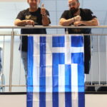 Two men stand behind a metal railing, one giving a thumbs-up, with a blue-and-white Greek flag hanging over the railing.