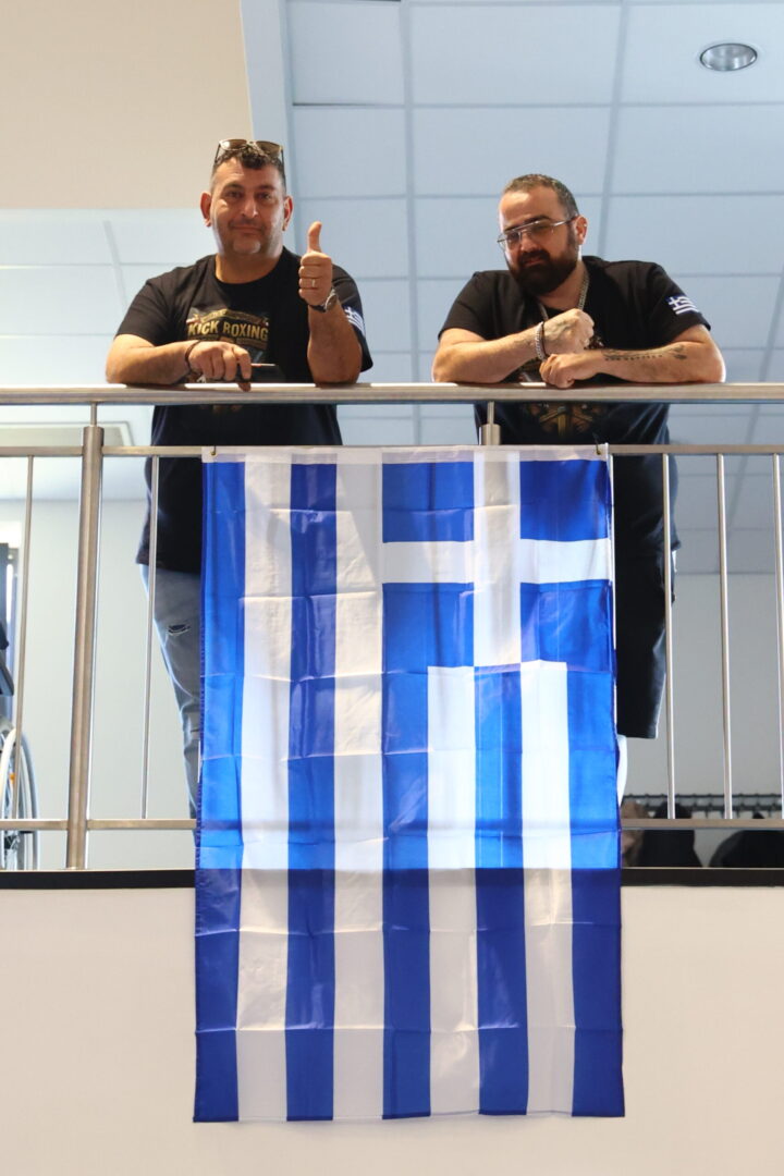 Two men stand behind a metal railing, one giving a thumbs-up, with a blue-and-white Greek flag hanging over the railing.