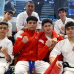 Group of smiling young martial artists posing together in white karategis and red warmups, seated in stadium seating and showing fists in a team photo