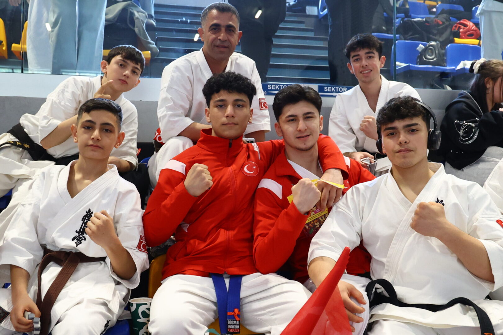 Group of smiling young martial artists posing together in white karategis and red warmups, seated in stadium seating and showing fists in a team photo