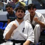 Three young martial artists wearing white gis pose for a photo in a venue, with a fist raised in the foreground and two teammates behind them walking onto the mat, spectators seated in the background.