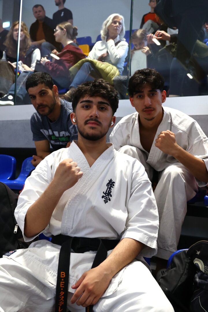 Three young martial artists wearing white gis pose for a photo in a venue, with a fist raised in the foreground and two teammates behind them walking onto the mat, spectators seated in the background.