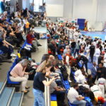 Crowded sports hall with spectators in blue seats watching a martial arts tournament in white uniforms on the mat far side.
