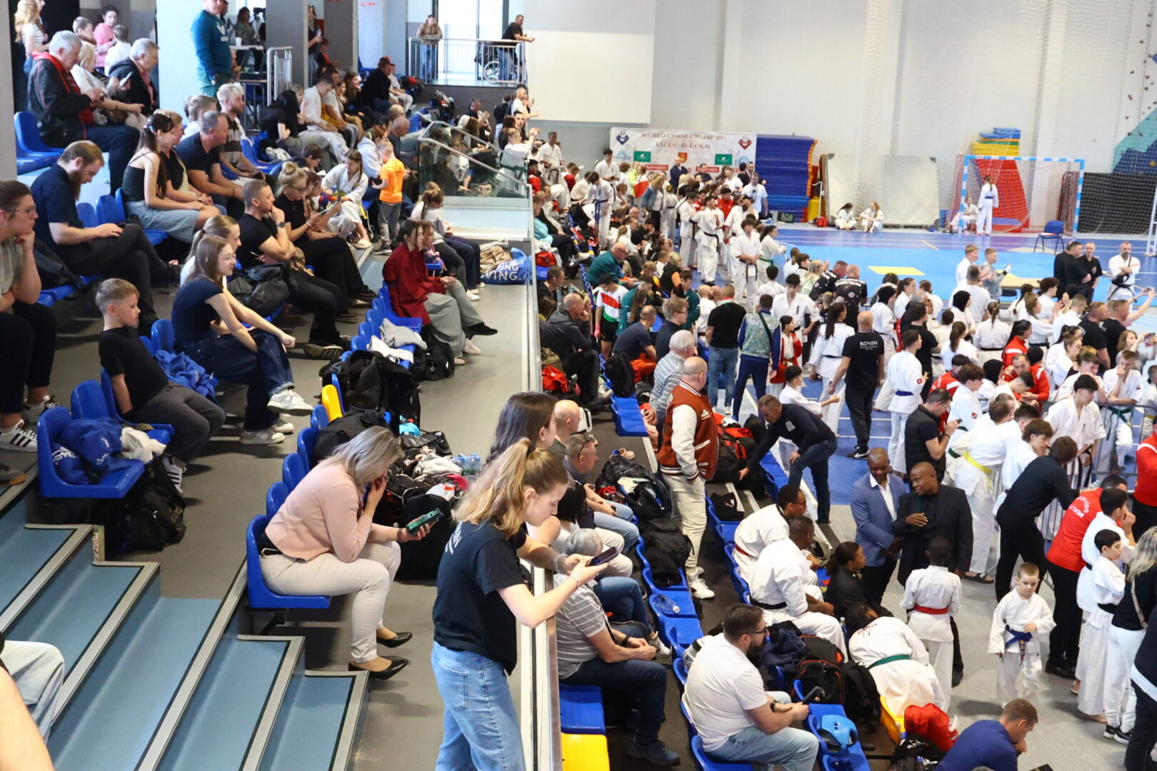 Crowded sports hall with spectators in blue seats watching a martial arts tournament in white uniforms on the mat far side.