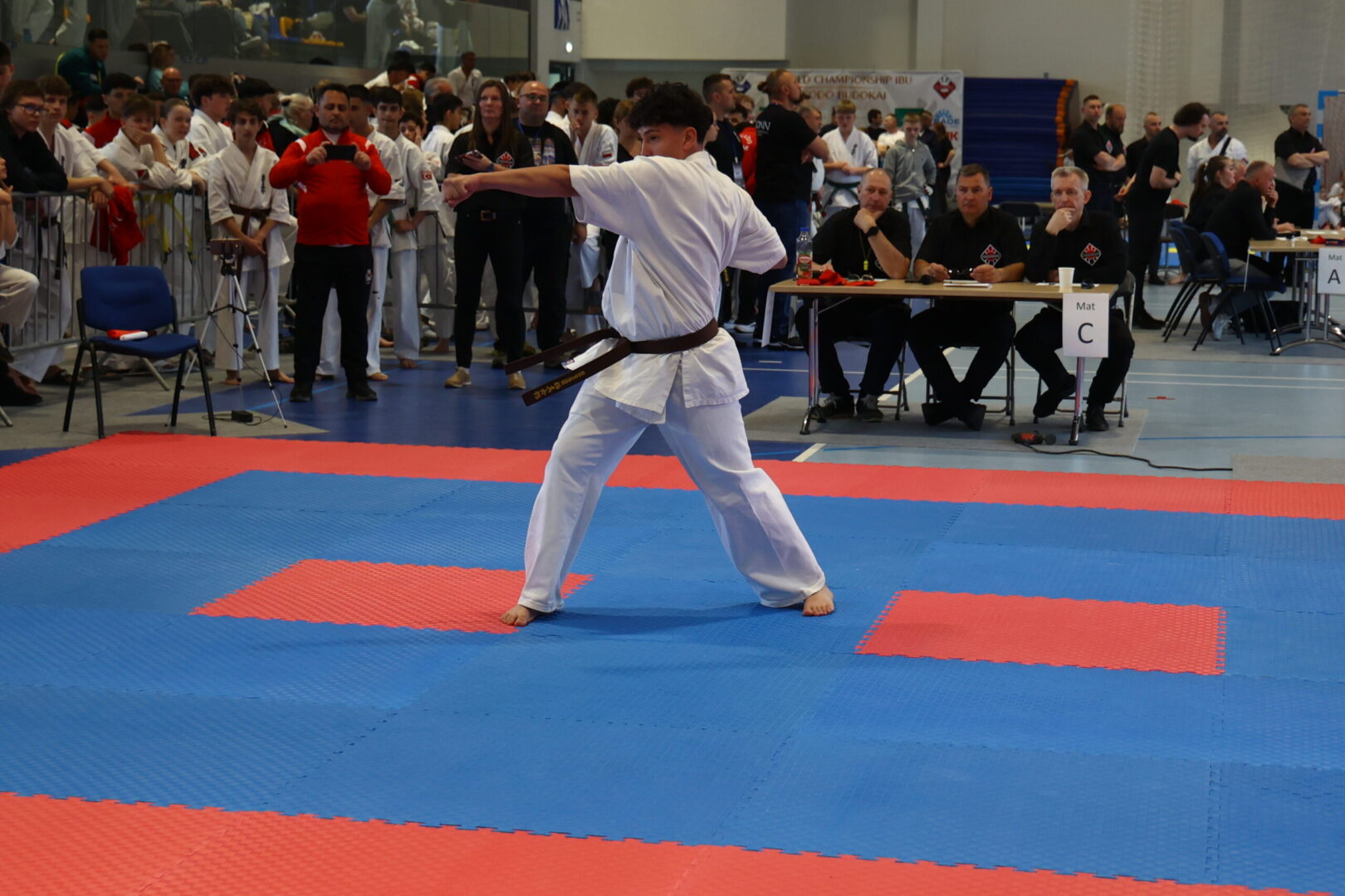 Karate competitor in a white gi and brown belt delivering a punch on blue-and-red competition mats, with judges and spectators in the background.