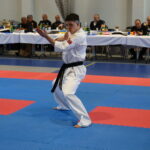 Karate competitor in a white gi with a black belt performs a kata on blue and red mats as judges watch from tables in the background.