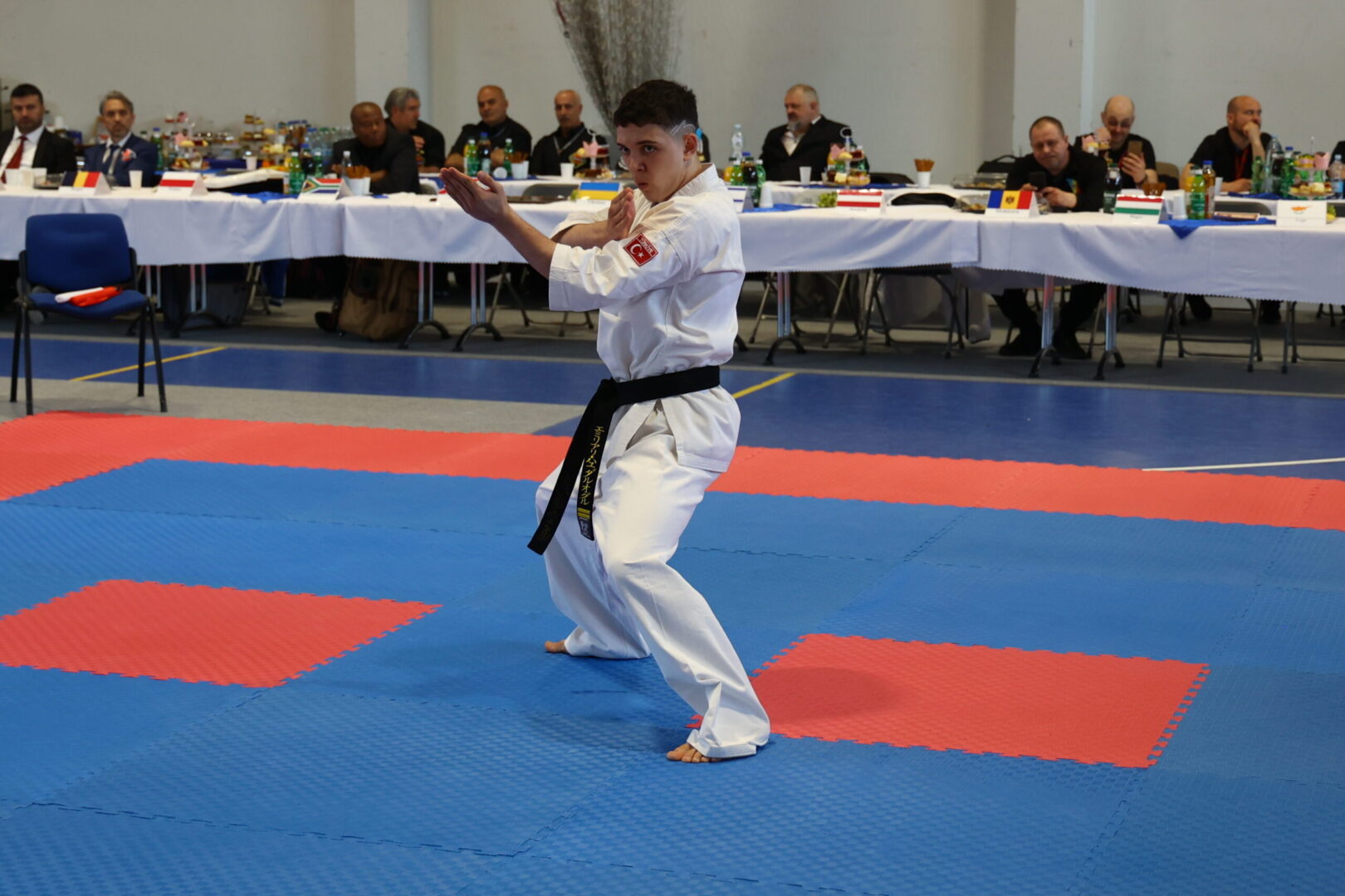 Karate competitor in a white gi with a black belt performs a kata on blue and red mats as judges watch from tables in the background.
