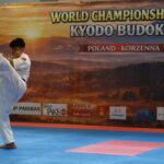 Karate athlete in a white gi delivering a high kick on blue-pink mats at a world championship venue.