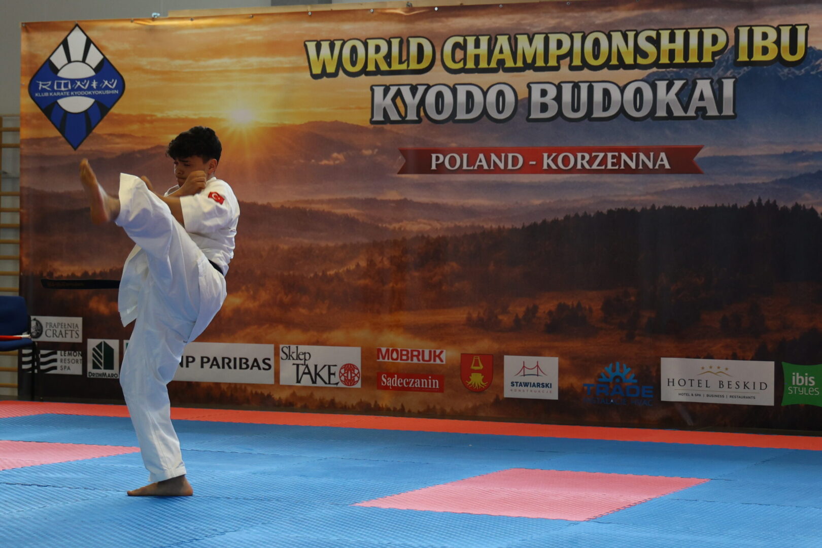 Karate athlete in a white gi delivering a high kick on blue-pink mats at a world championship venue.