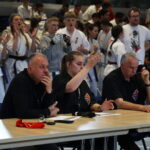 Three judges sit at a table scoring a martial arts event, with a crowd of competitors in white gis behind them.