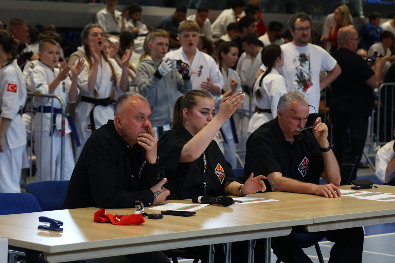 Three judges sit at a table scoring a martial arts event, with a crowd of competitors in white gis behind them.
