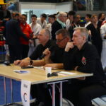 Three men in black shirts sit at a judging table for a karate competition, with competitors in white gi behind them.