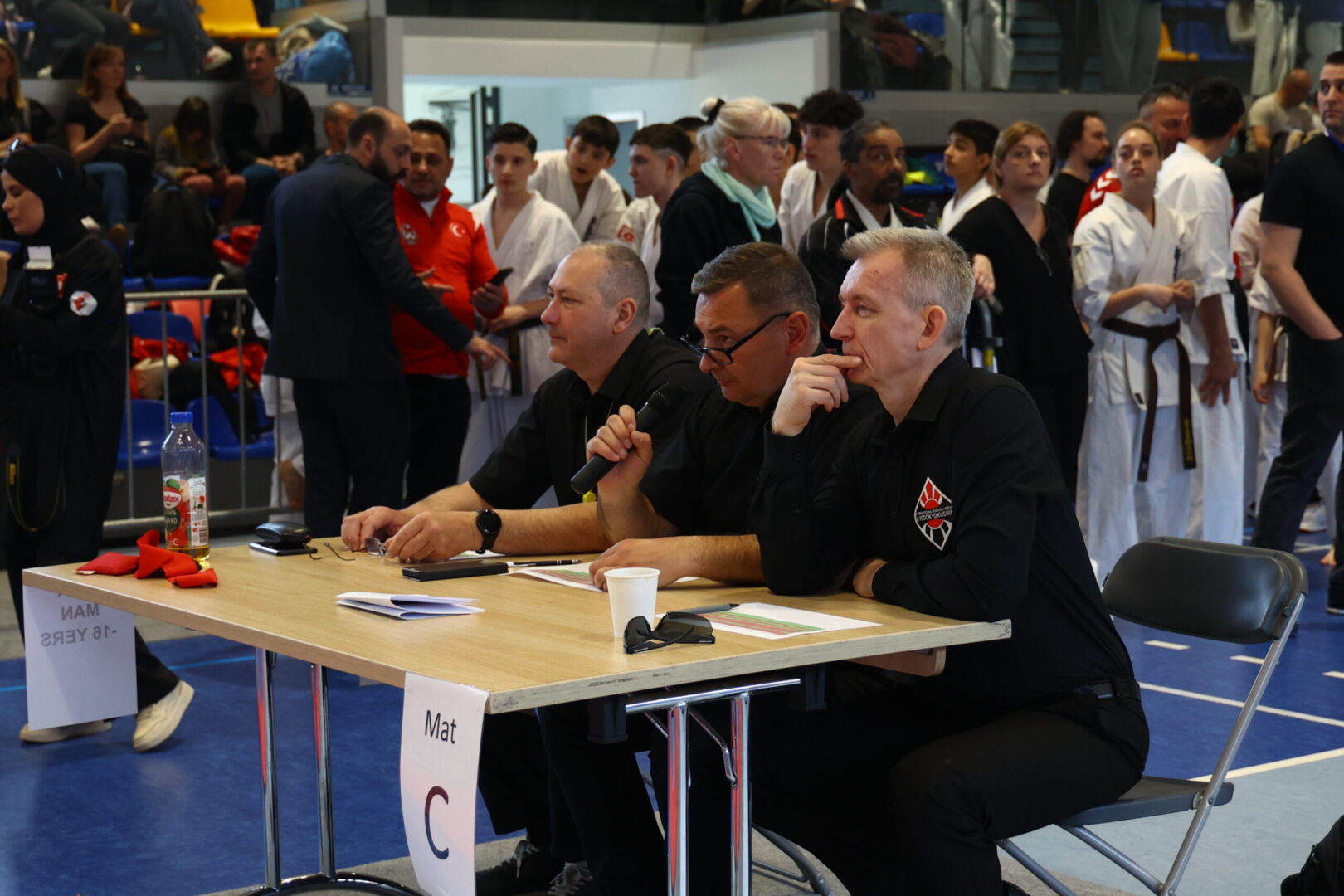 Three men in black shirts sit at a judging table for a karate competition, with competitors in white gi behind them.
