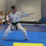 Woman in a white karate gi with a black belt performs a kata, extending her right arm in a wide stance on blue mats, with teammates watching in the background.