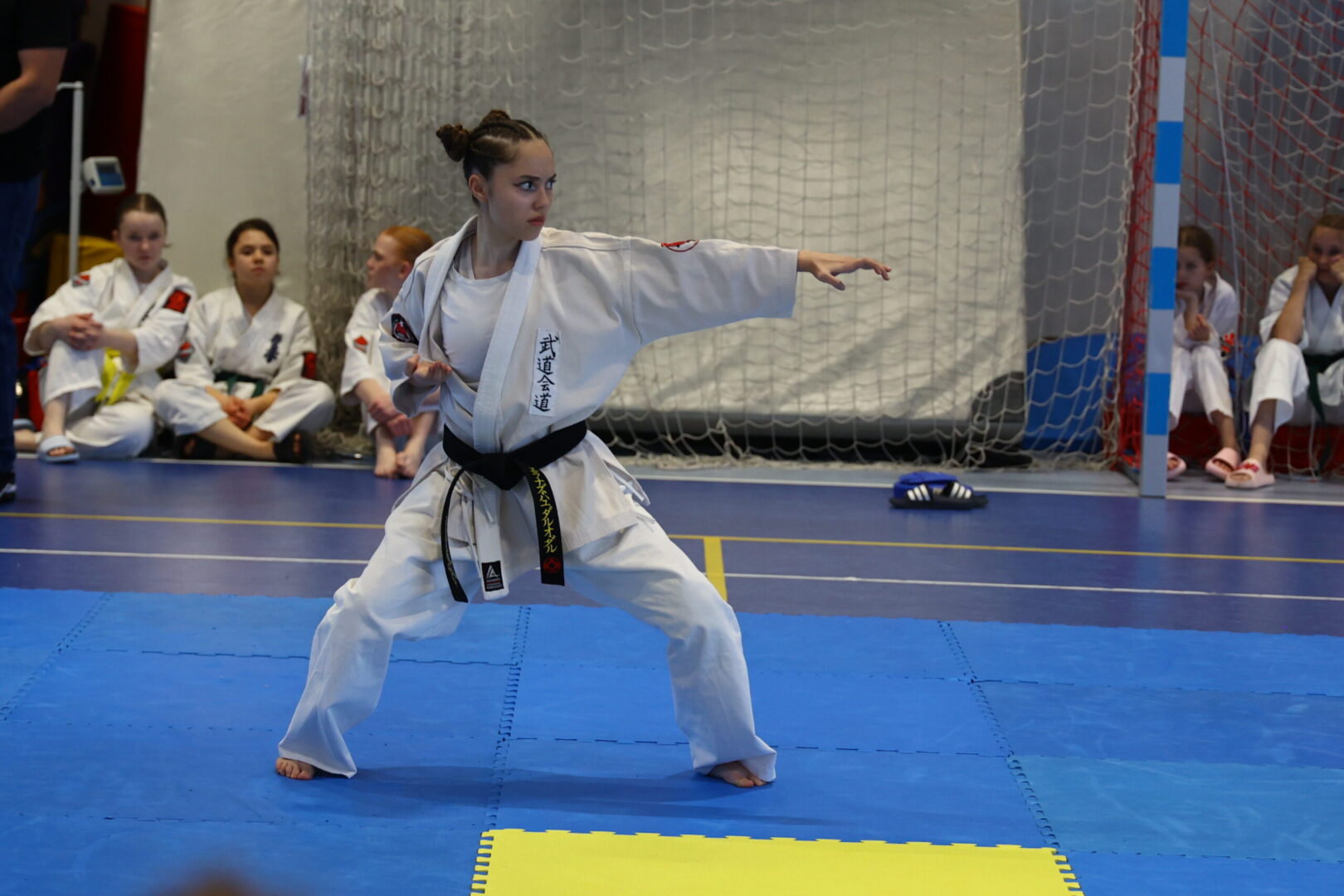 Woman in a white karate gi with a black belt performs a kata, extending her right arm in a wide stance on blue mats, with teammates watching in the background.