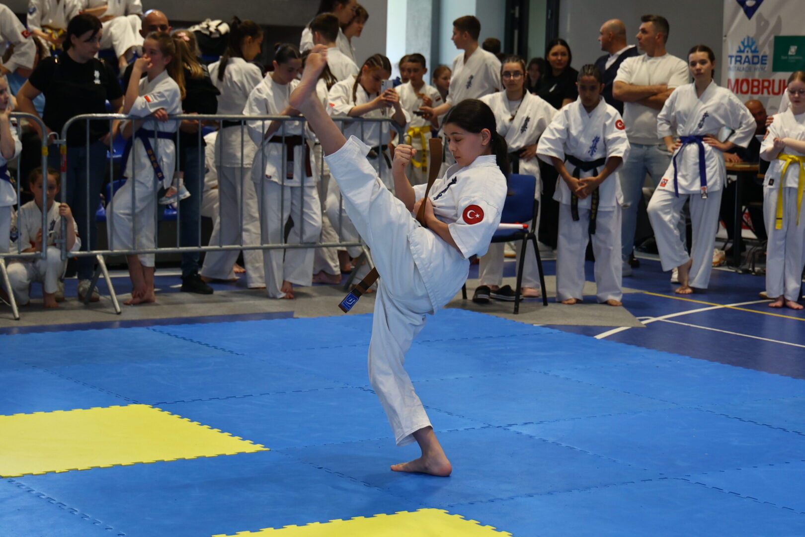 Young girl in a karate gi performs a high kick during a tournament, with spectators in white uniforms watching behind a barrier.
