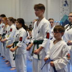 Group of young martial artists in white gi with green belts lined up on blue mats, indoors for a ceremony or training session.